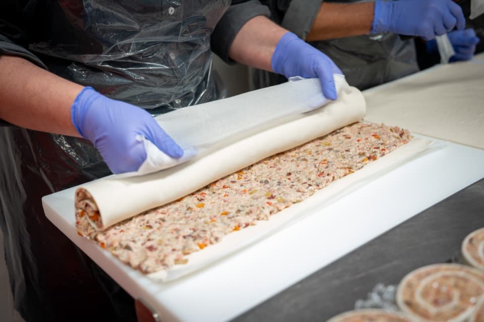 Hände in blauen Handschuhen rollen eine Füllung in Teig – Handarbeit in der Gastroabteilung.
